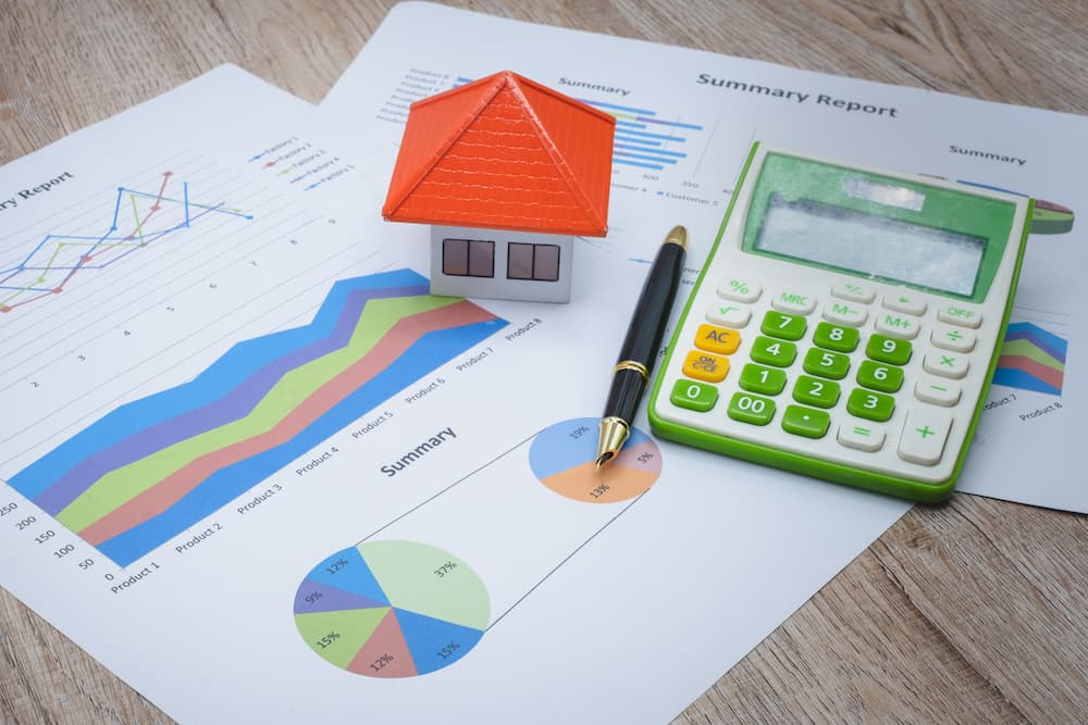 A model home and a calculator sit on top of scattered papers with graphs and info graphics regarding investing in rental properties